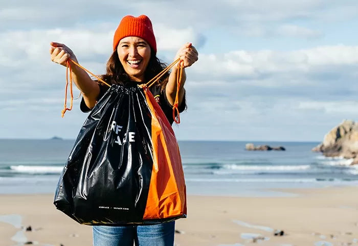 A girl standing on the beach, holding a SRFACE bag designed to store a wetsuit, with the bag’s logo visible
