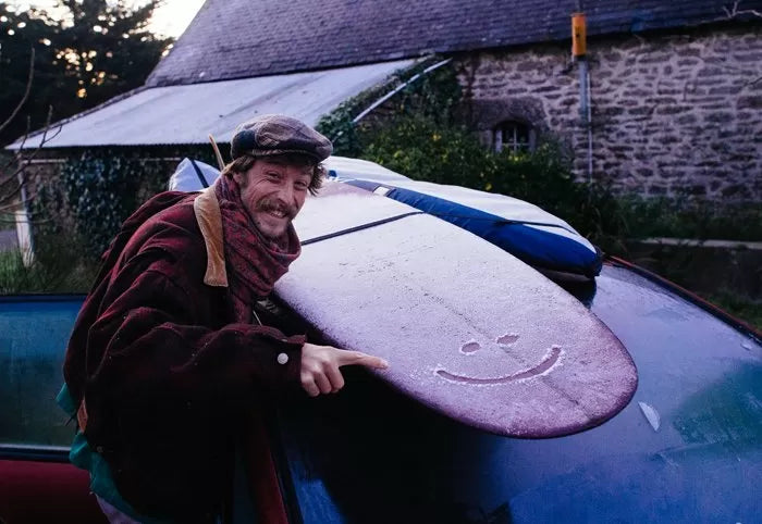 A happy surfer standing next to his car on a cold winter day, smiling as he holds his longboard