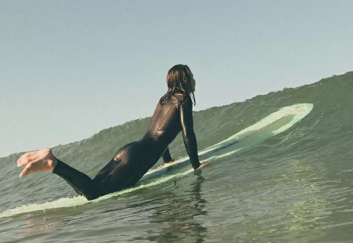 A female surfer in a SRFACE Flex wetsuit, laying on her longboard and waiting for the wave to pass