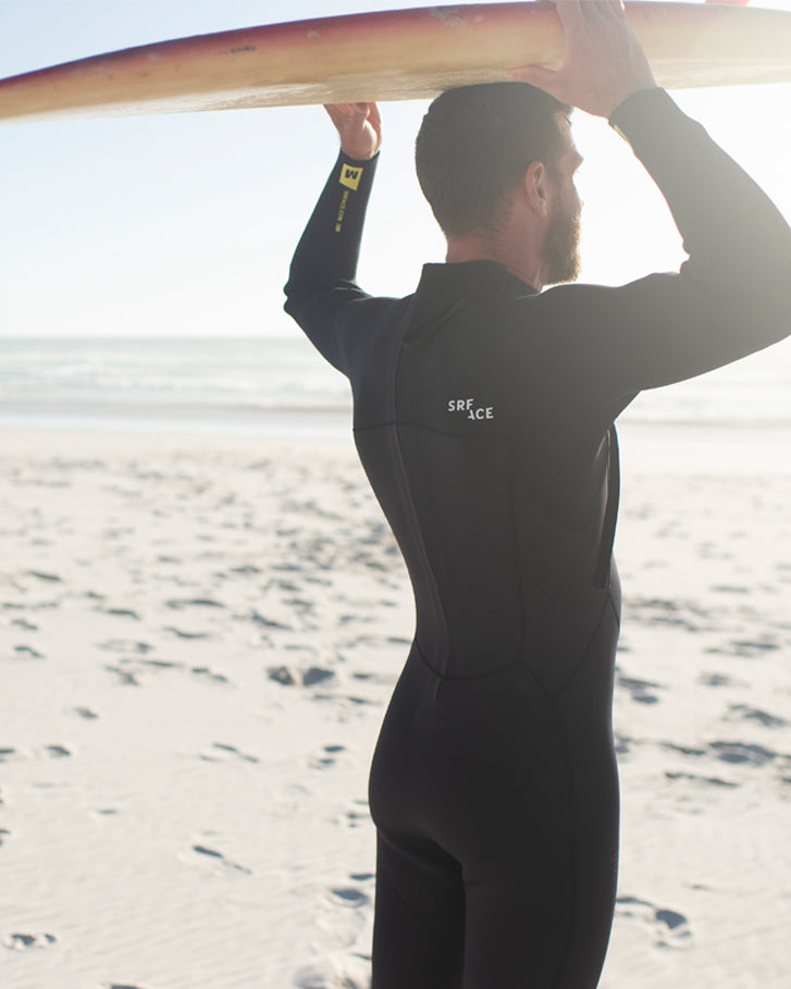 A beginner surfer in a SRFACE school wetsuit, standing on the beach with their surfboard, ready to hit the water