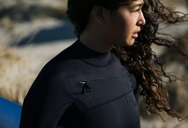 A portrait of a girl wearing an SRFACE Eco wetsuit on the beach, standing with her face turned toward the sun