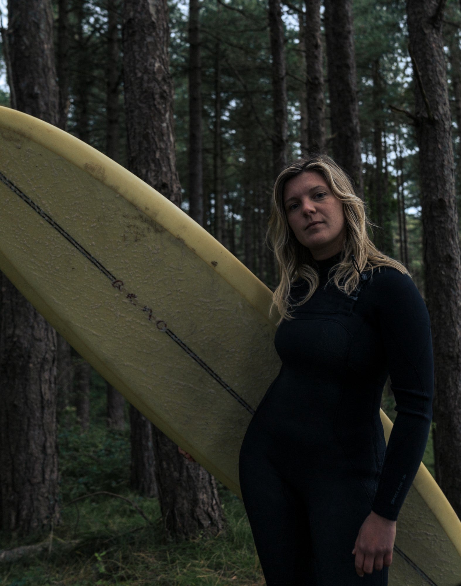 Laura poses for the camera, longboard in hand, wearing her SRFACE wetsuit