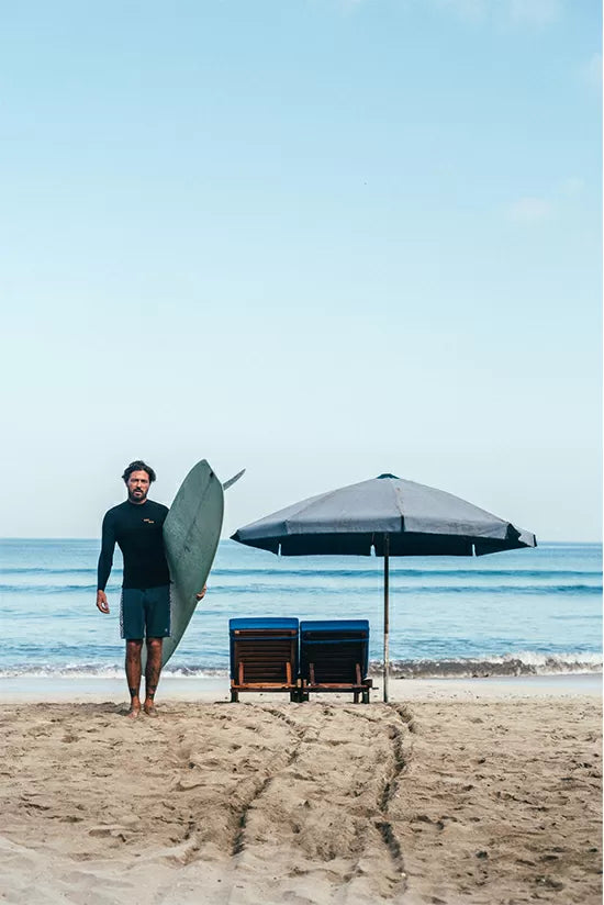 Raskal standing on the beach next to a sunbed with his longboard