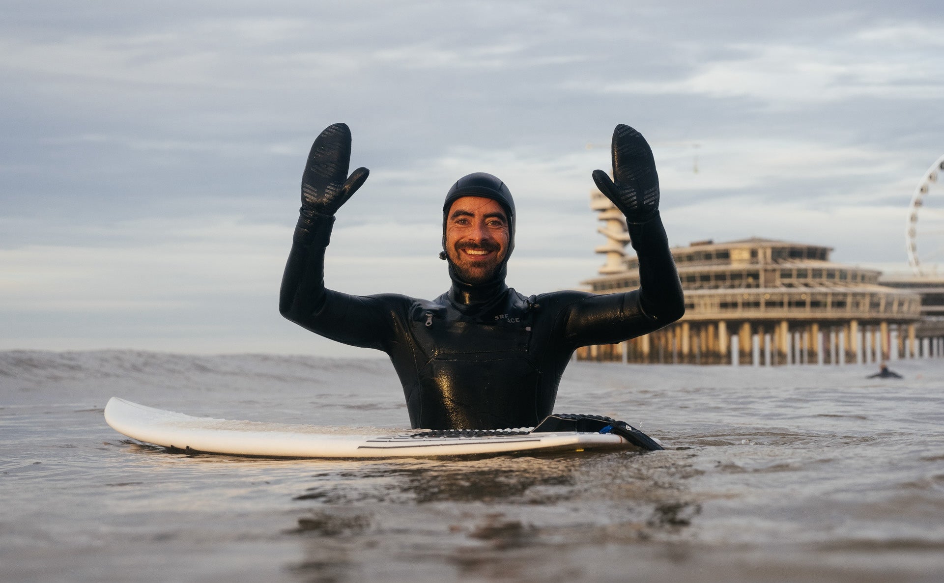 A happy surfer wearing a hooded SRFACE wetsuit and mittens, floating in the water with his surfboard in front of him